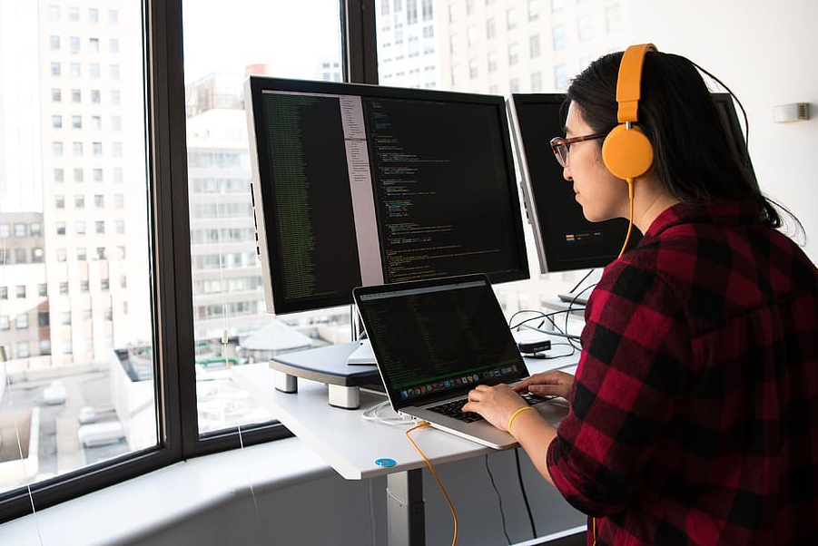 a woman in yellow headphones sits in front of a laptop and monitors with lines of code visible